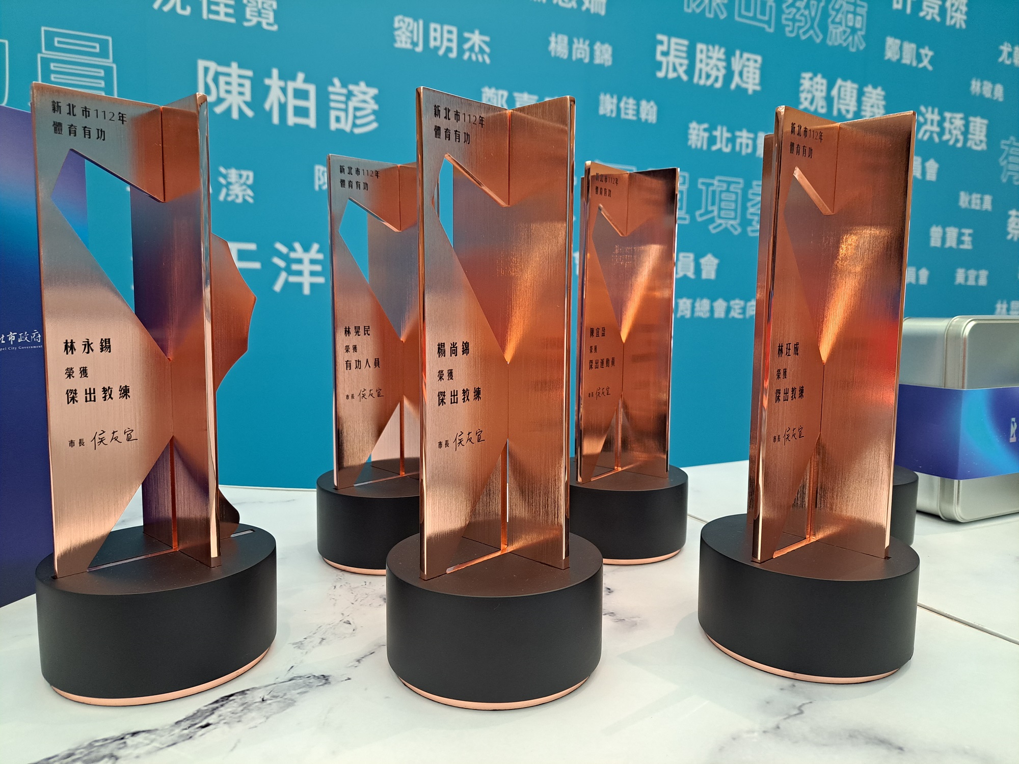 新北市112年體育有功質感女王頭獎座，以虛實交織與鏡面的十字反射手法轉化呈現，搭配玫瑰金色的女王頭，象徵淬鍊過程的璀璨火紅